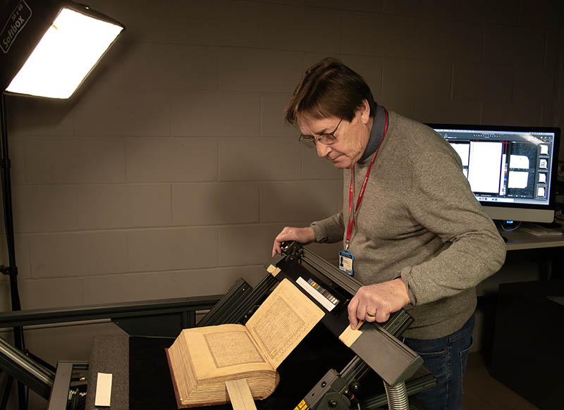 A man stands over a conservation cradle holding an old book, with a computer monitor in the background