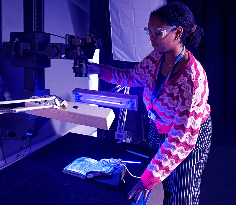 A woman adjusts an overhead camera; ultraviolet light is shining on a book in a cradle
