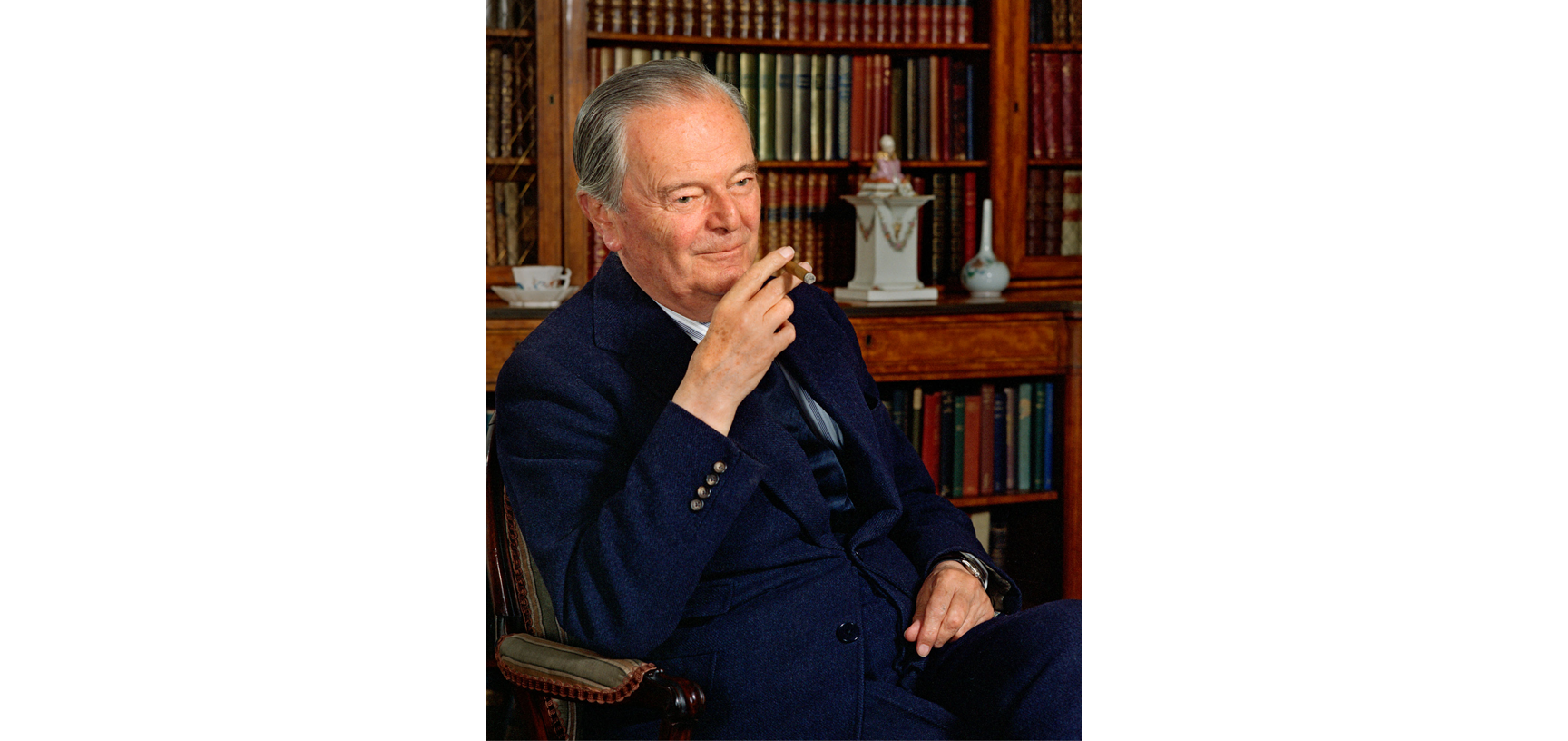 Portrait of Kenneth Clark - an older white male with grey hair, dressed in a navy blue suit, blue stripey shirt and navy tie, sitting in a wooden chair looking diagonal to the camera with bookshelves in the background and a cigar in his hand