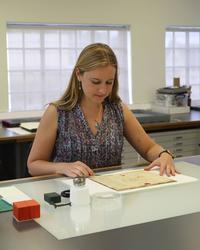 A woman stands next to a desk, she has a manuscript in front of her - to the left are glass bottles and other conservation equipment