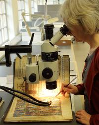 Julia Bearman A woman looks through a microscope, beneath the microscope is an open book. The woman is using a paint brush to carry out conservation work on it.